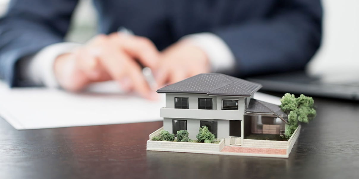 テーブルの上に置かれた戸建て住宅の模型と、背景で書類に記入するスーツ姿の人物